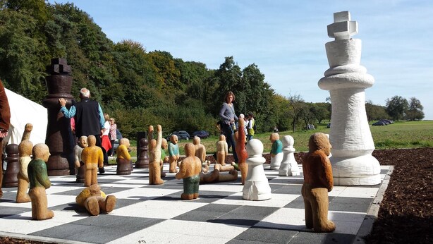 Ein großes Schachbrett im Freien mit übergroßen Schachfiguren, umgeben von Menschen und Bäumen.