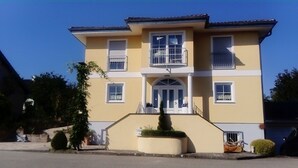 Ein zweistöckiges Gebäude mit einer symmetrischen Fassade, einem zentralen Eingang mit einem kleinen Vorbau und einem Balkon im oberen Stockwerk.