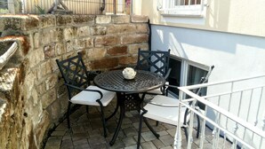 Eine kleine Terrasse im Freien mit einem runden Metalltisch und zwei Stühlen, jeweils mit einem weißen Sitzkissen, sowie einer dekorativen Vase auf dem Tisch.