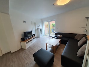 Ein modernes Wohnzimmer mit einem Flachbildfernseher auf einem Holzständer, einem schwarzen Sofa, einem Holztisch und einem schwarzen Hocker.