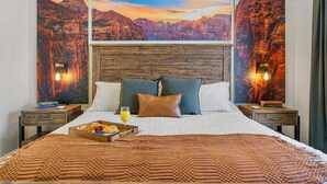 Une chambre d’hôtel avec un grand lit, une tête de lit en bois, deux tables de chevet, un plateau avec le petit-déjeuner et, au mur, une vue sur le coucher de soleil.