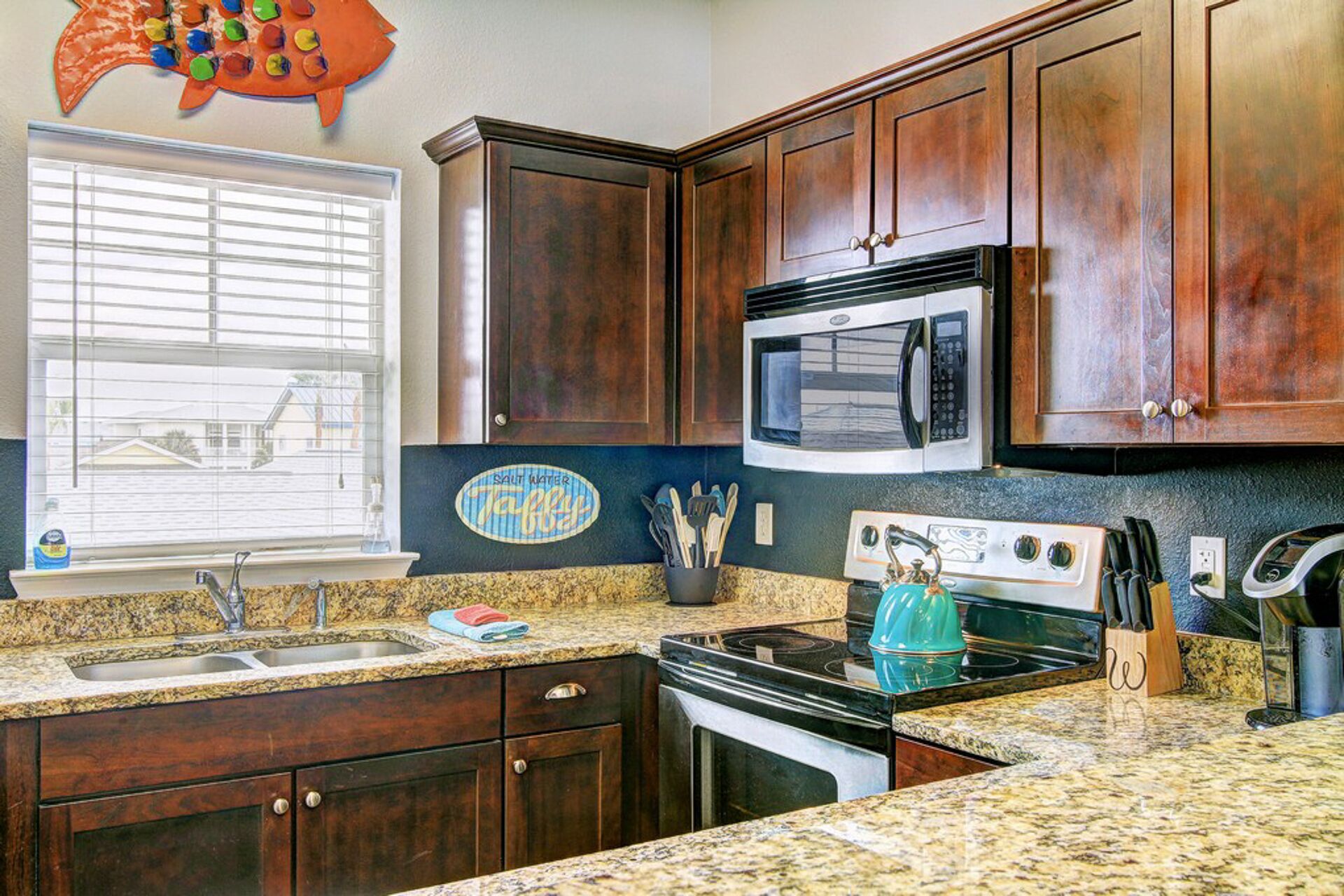 A kitchen with dark wooden cabinets, a granite countertop, a stove, an oven, a microwave, and a window with blinds.
