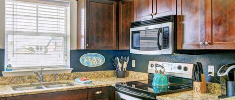 A kitchen with dark wooden cabinets, a granite countertop, a stove, an oven, a microwave, and a window with blinds.