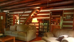 A cozy log cabin interior with a wooden staircase, two sofas, a coffee table, and a bookshelf filled with books. - River’s Edge Cabin- Perfect for Romance/Outdoor Adventures/Relaxation (Elkins)