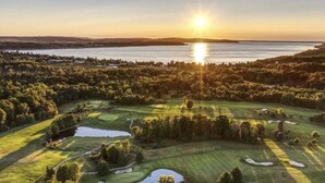 A golf course with a lake, surrounded by dense forest, and a sunset in the background.