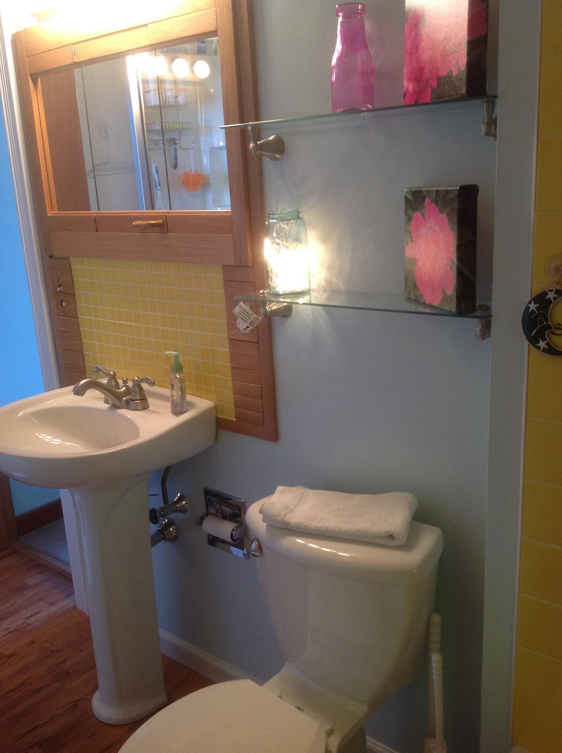 A bathroom with a white sink, toilet, and a mirror above the sink. There are shelves with decorative items and a glass jar with a light.