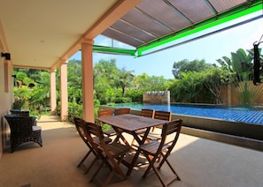 Uma área externa coberta com mesa e cadeiras de madeira, piscina e vegetação exuberante ao fundo.