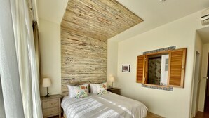 A bedroom with a wooden ceiling, a bed with floral pillows, a nightstand with a lamp, and a mirror with a decorative frame. - Spoondrift Hostal (Hengchun)
