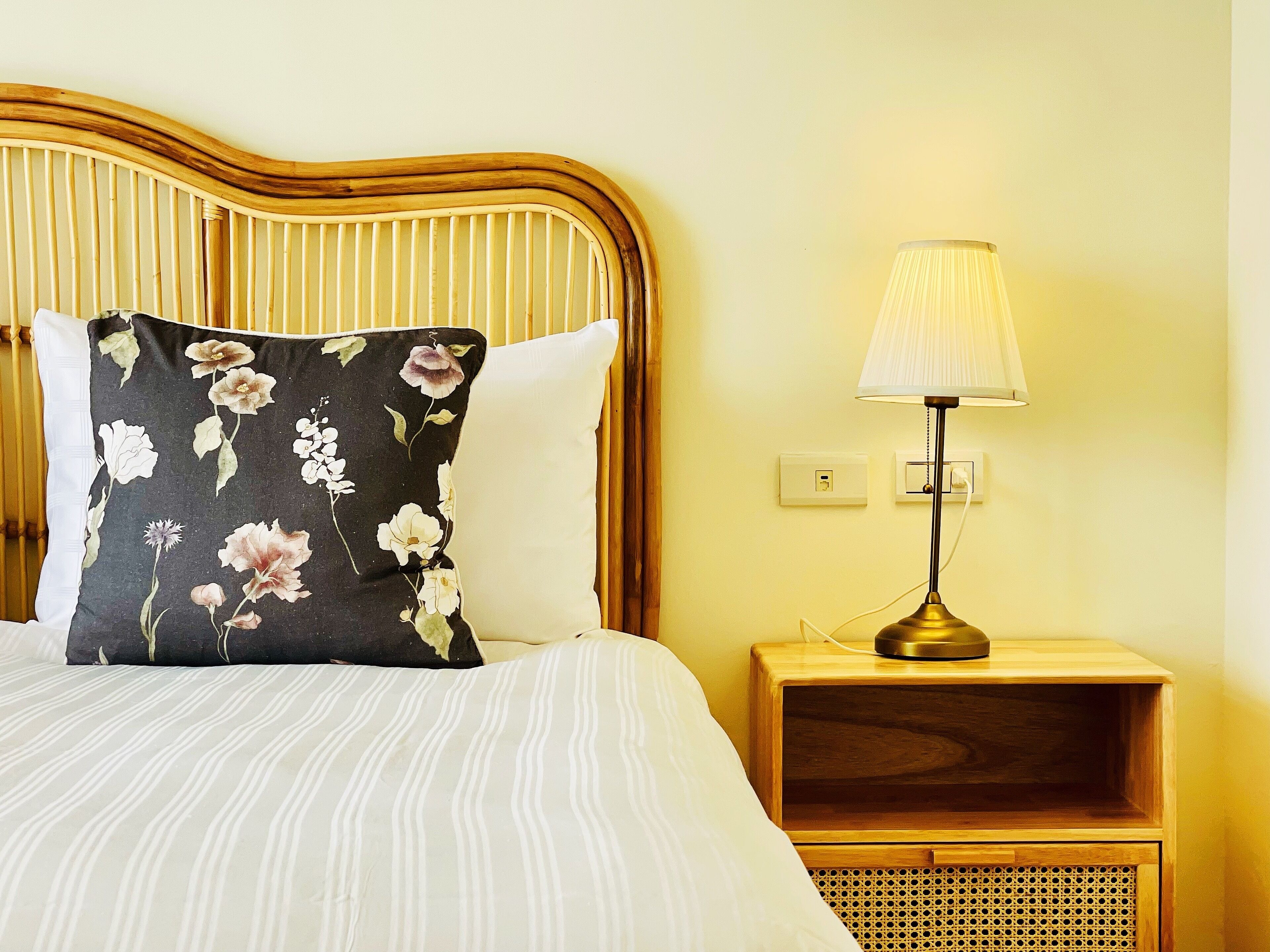 A bed with striped bedding, a floral pillow, a bedside lamp, and a wooden nightstand with a woven basket.