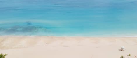 En strand med fri utsikt över havet, en sandstrand och en liten byggnad omgiven av grönska.