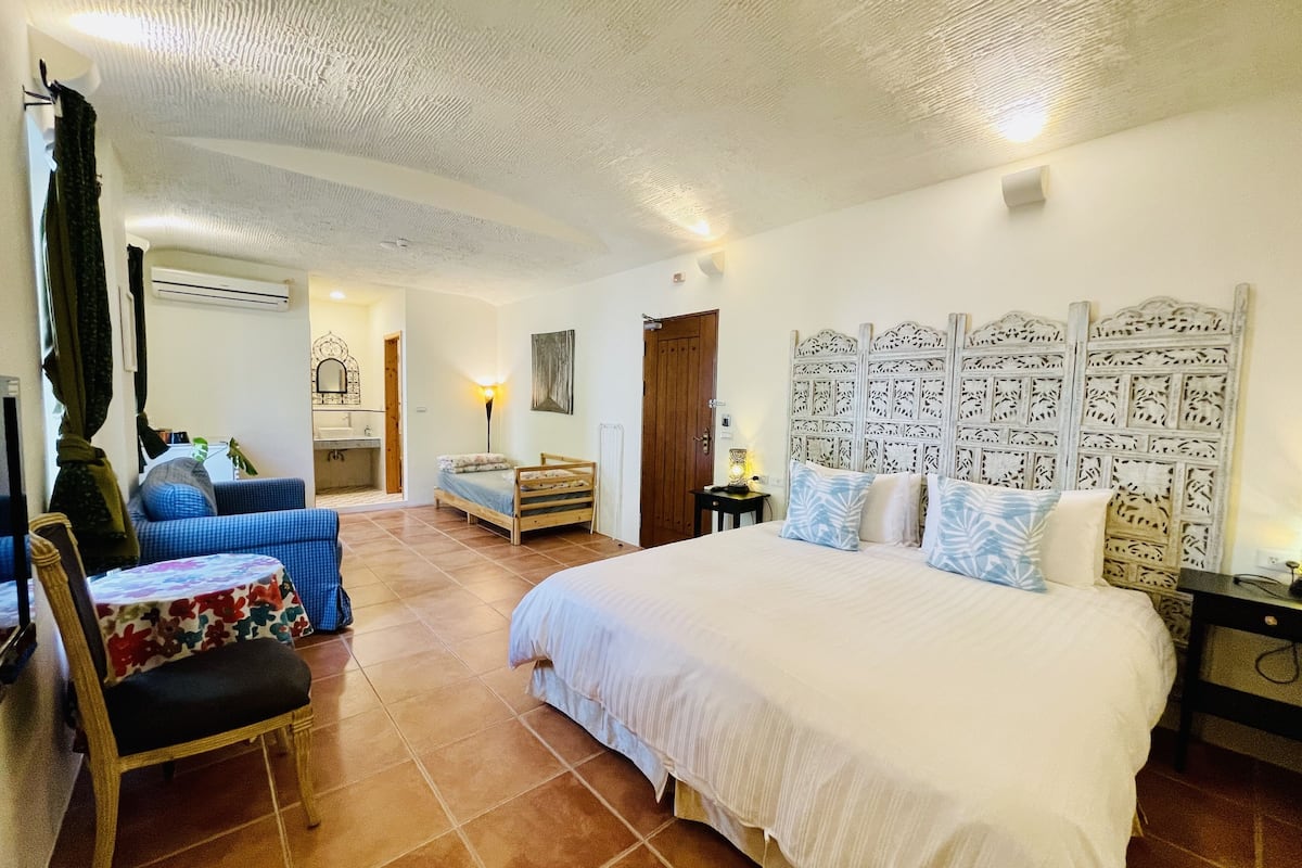 Un dormitorio con una cama grande, una silla, una mesita, un sofá y un baño al fondo.
