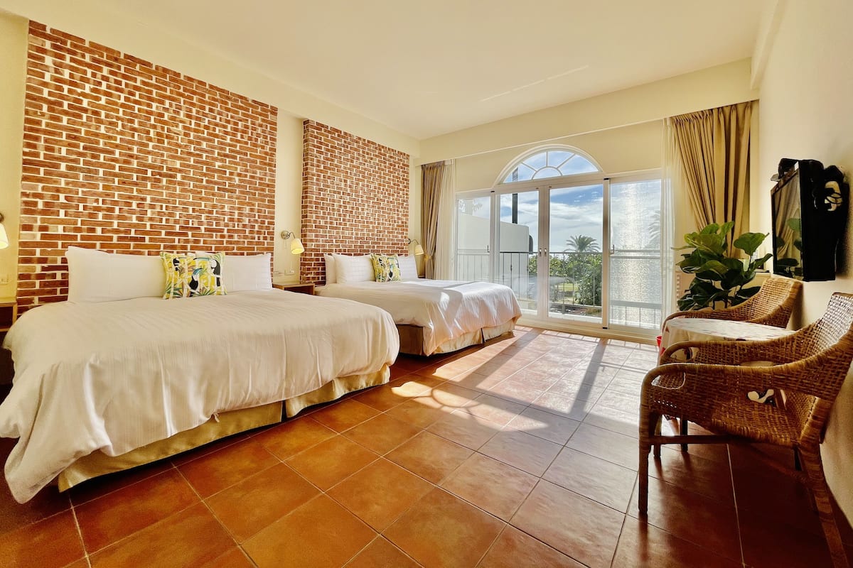 Habitación de hotel con dos camas, una pared de ladrillo, un balcón con vistas y una silla de mimbre.