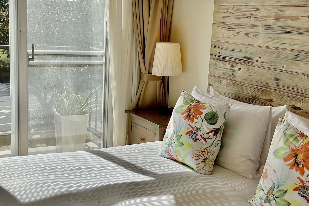Una habitación de hotel con cama, ventana con cortinas, mesita de noche y una lámpara, y un cojín estampado con motivos florales.