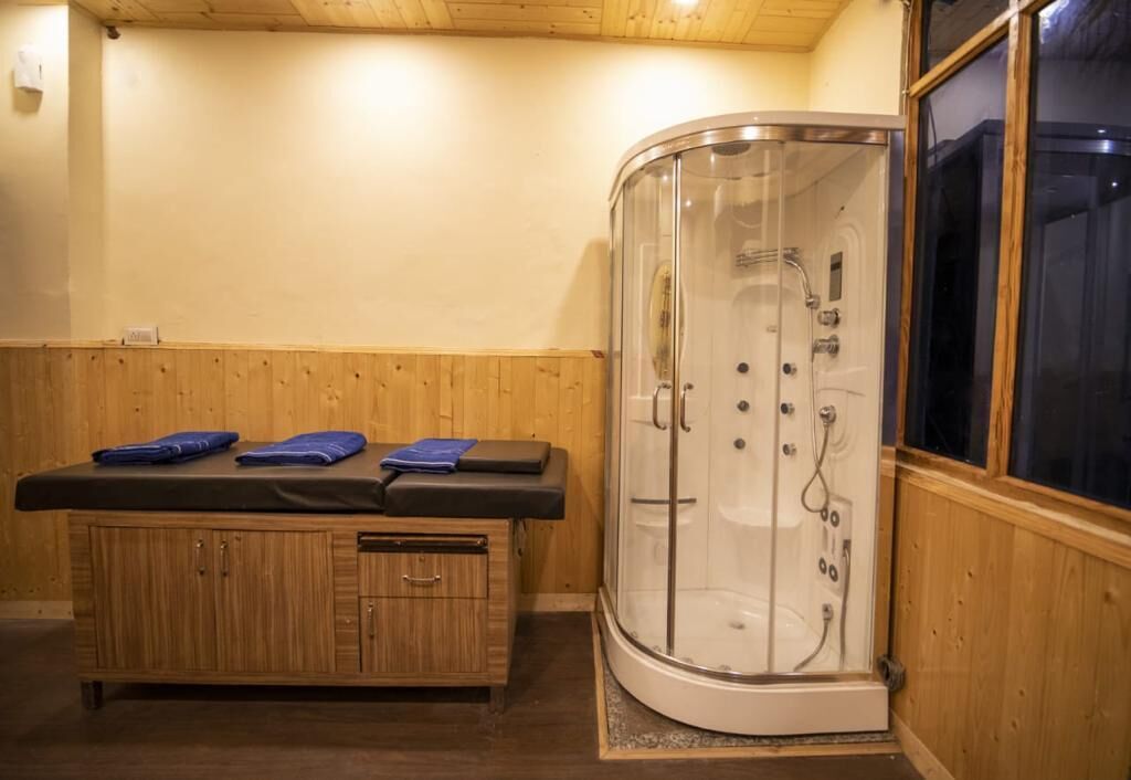 Un mueble de madera con encimera negra y toallas azules, una zona de ducha con cristal y suelo de madera, y una ventana con vistas oscuras.