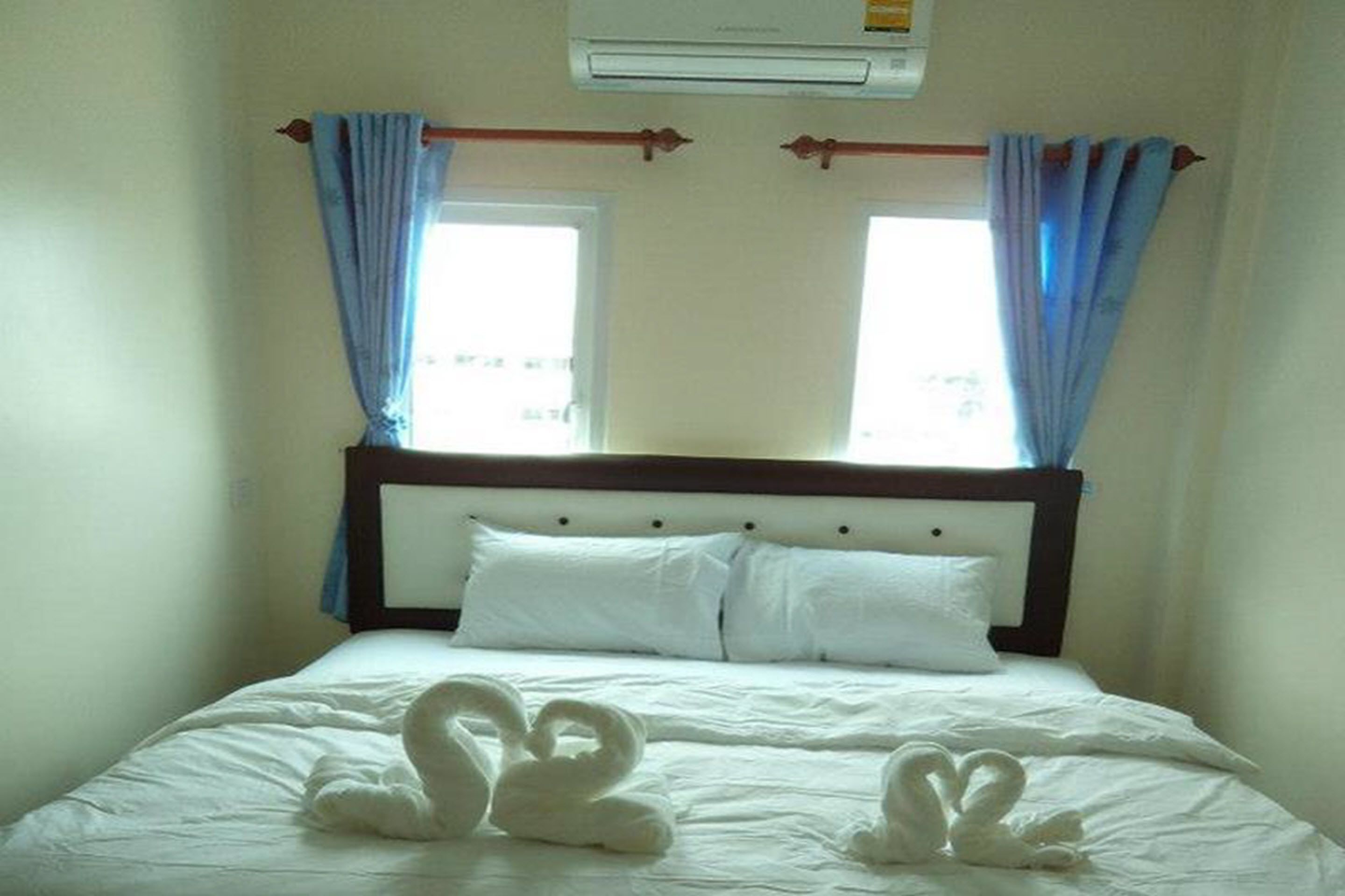 A hotel room with a bed, white linens, a wooden headboard, two windows with blue curtains, and a wall-mounted air conditioner.