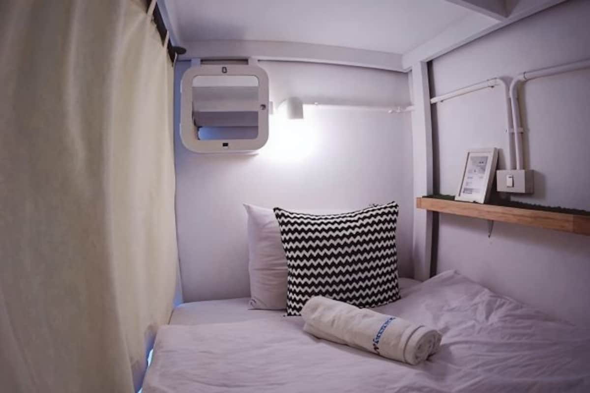 Una cabaña pequeña y privada con una cama, un almohadón, una manta, una repisa con una foto y una ventana con cortinas.