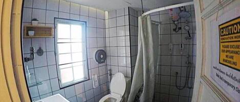 Une salle de bain comprenant des toilettes, un lavabo et un espace douche. Un panneau d’avertissement indique que des travaux de rénovation sont en cours.