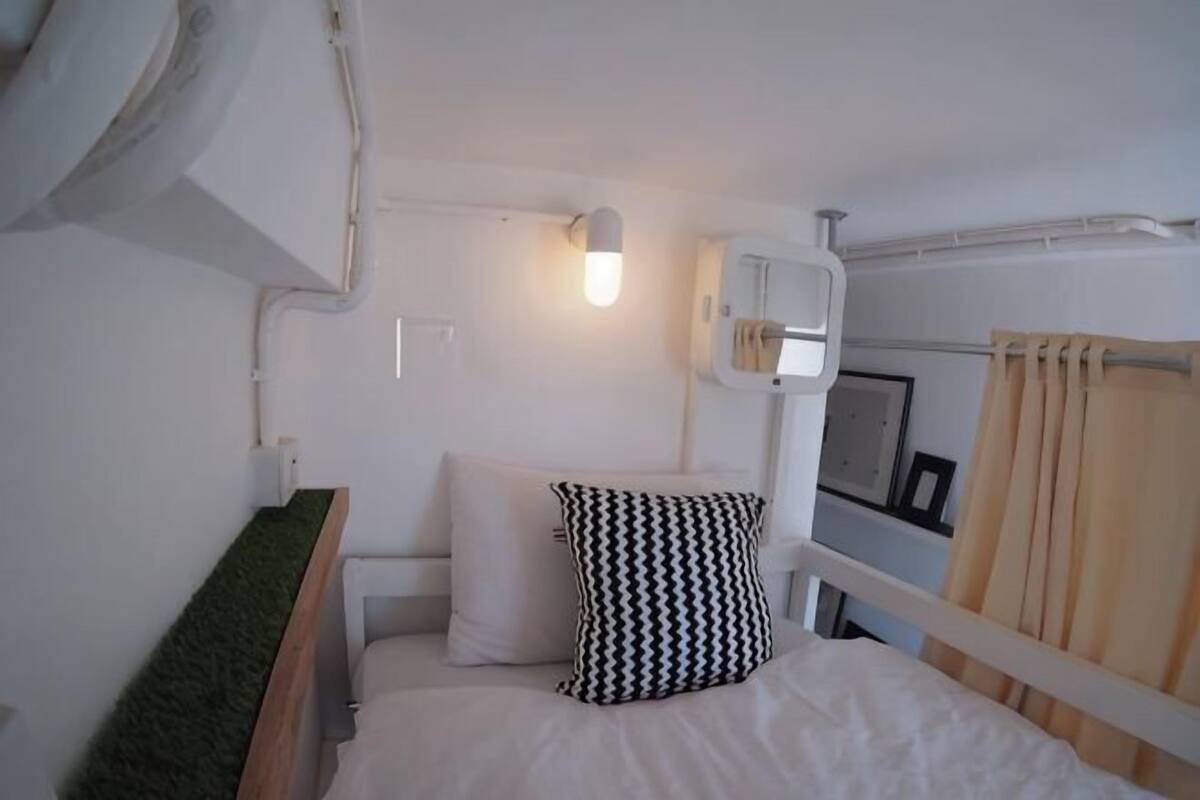 A small, single-bed room with a white headboard, a bedside lamp, a wall-mounted air conditioning unit, and a curtain.