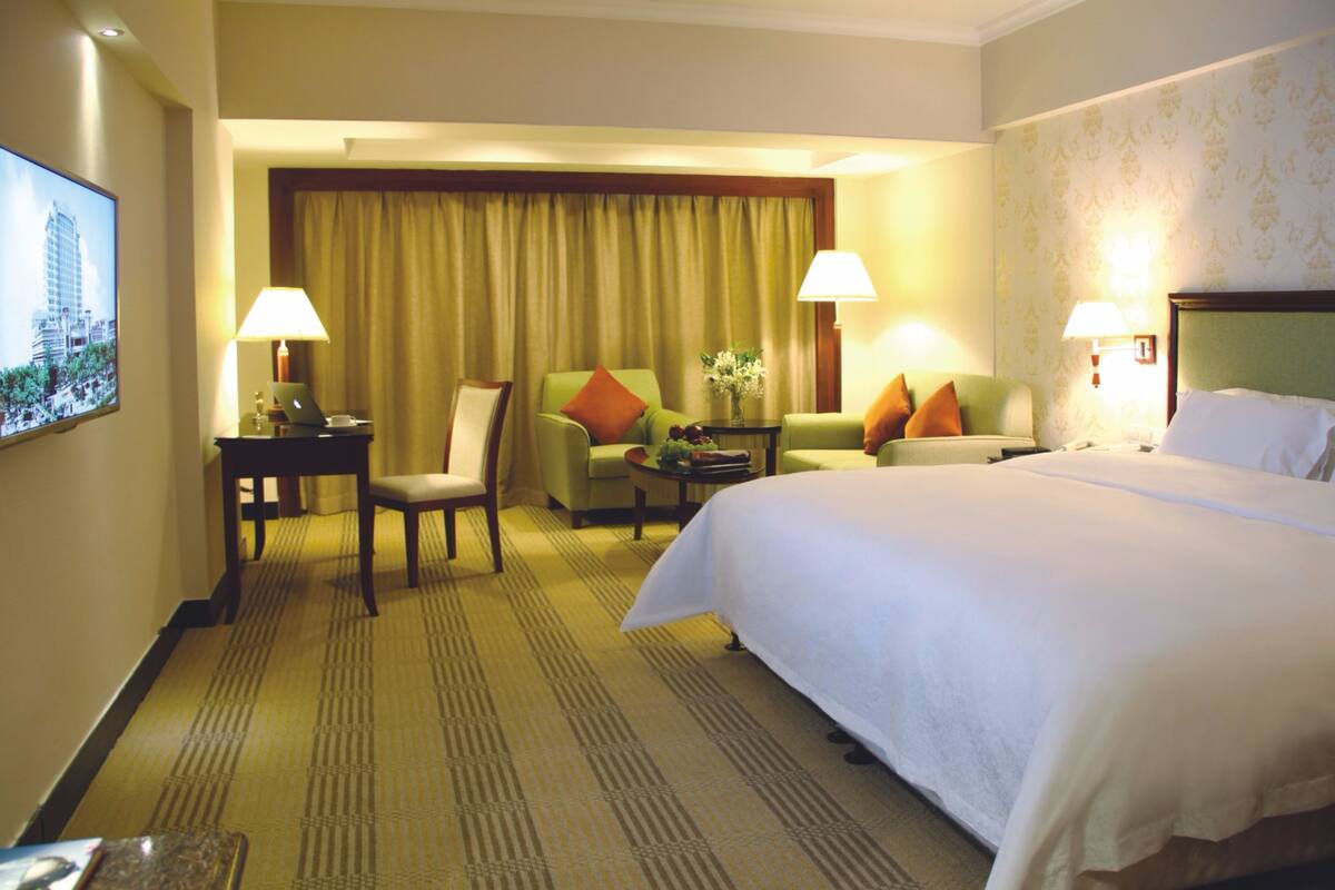 A hotel room with a large bed, two armchairs, a desk with a laptop, and a television on the wall.