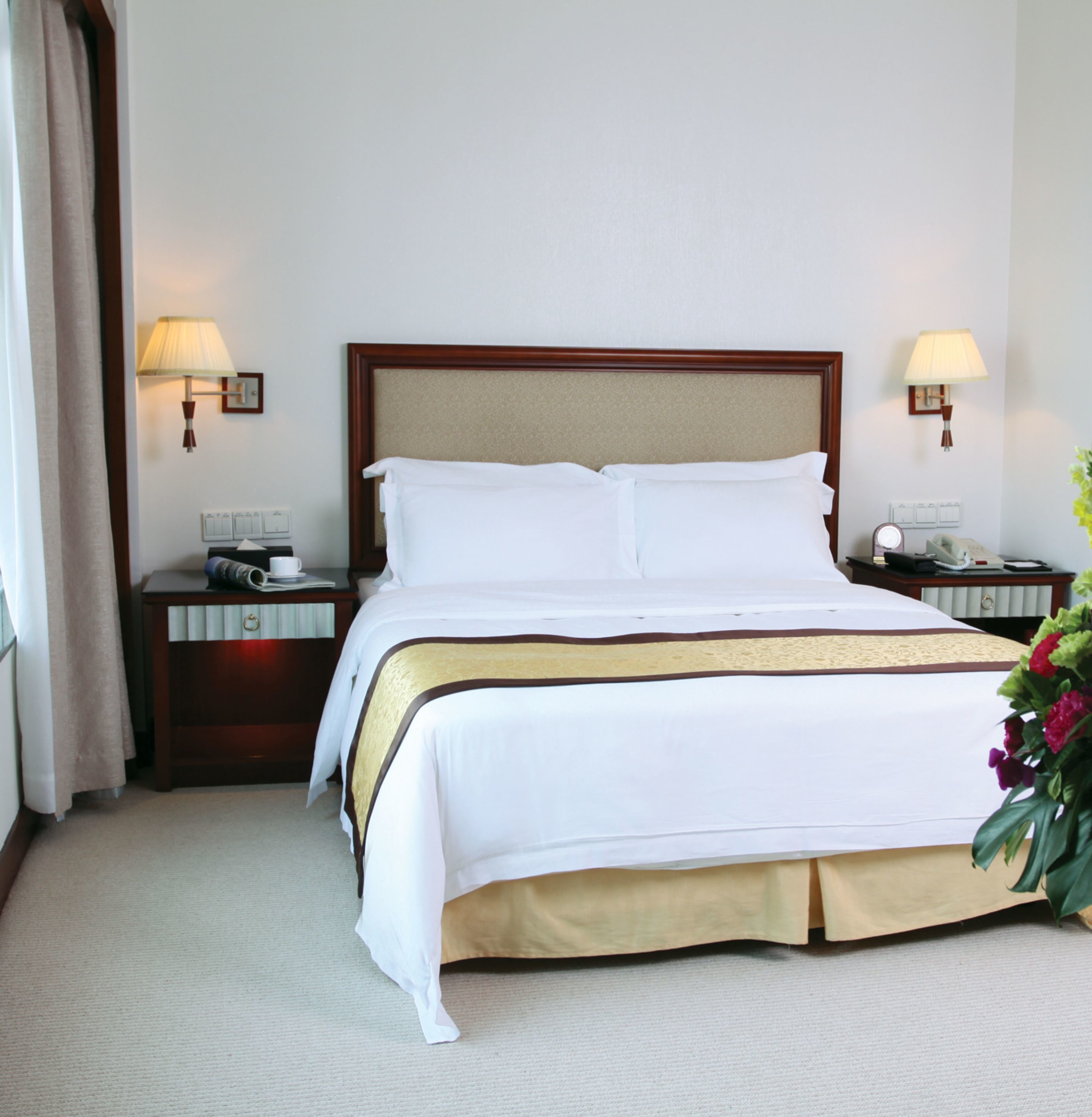 A hotel room with a large bed, two bedside tables, a nightstand, a lamp, and a flower arrangement.