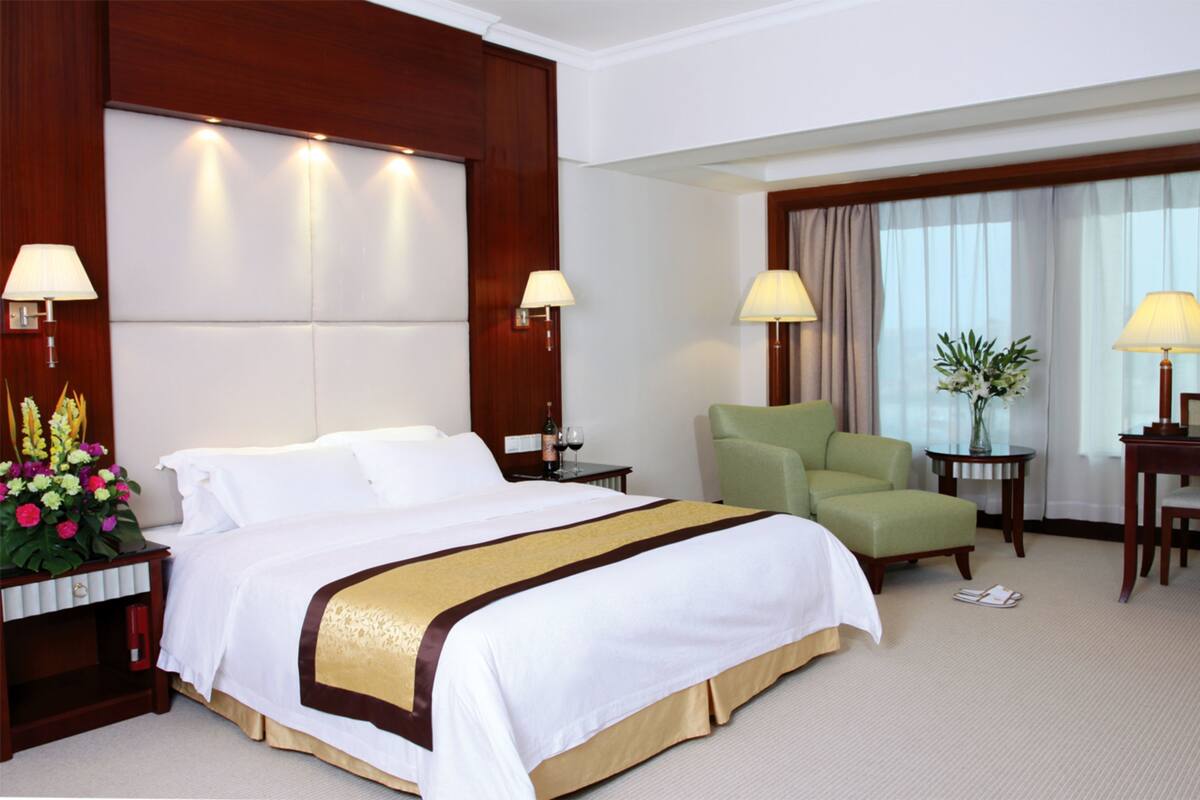 A hotel room with a large bed, two bedside tables, a green armchair, a small table with a vase, and a window with curtains.