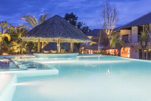 Une piscine agrémentée d’un gazebo à toit de chaume, de palmiers et d’un bâtiment doté d’un balcon.