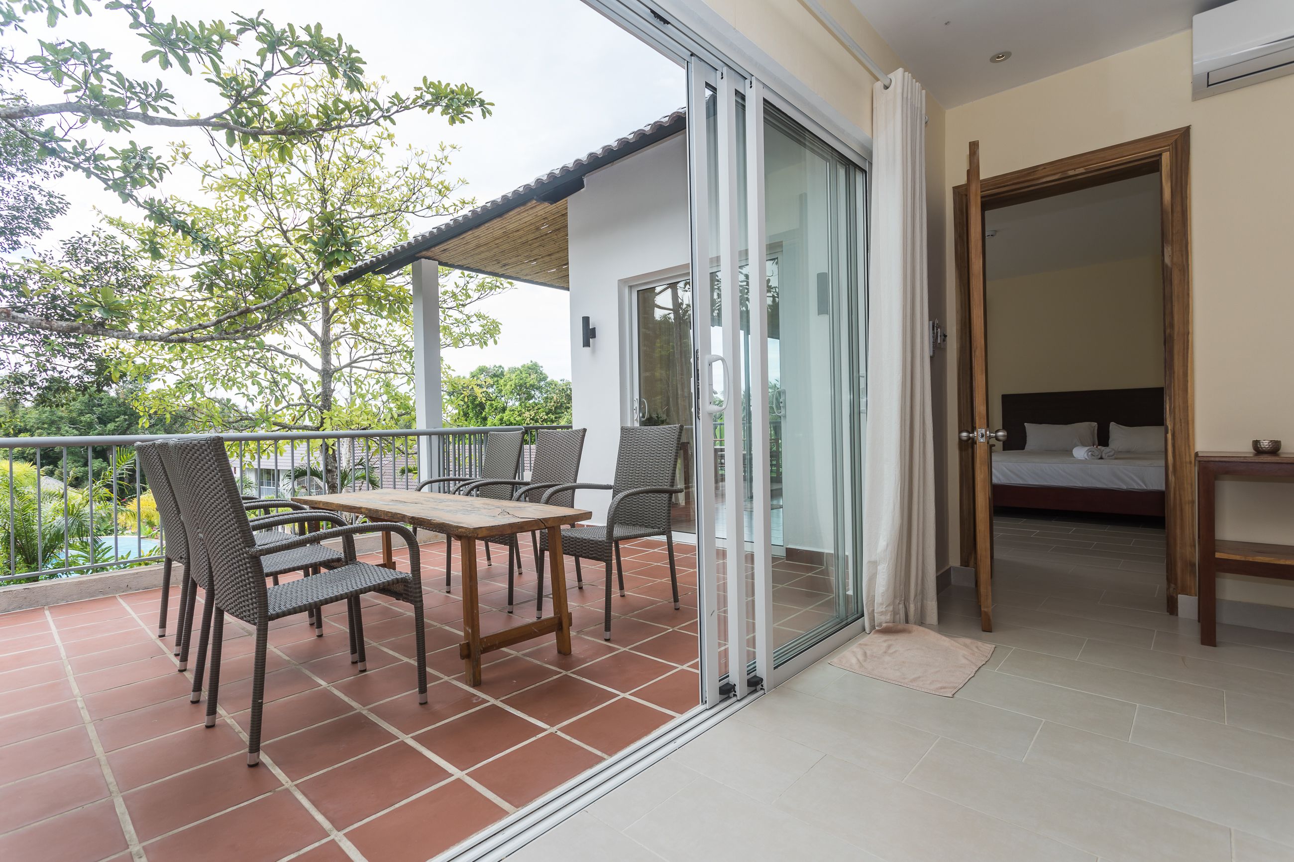 A balcony with a table and chairs, a bed in the adjacent room, and a view of greenery outside.