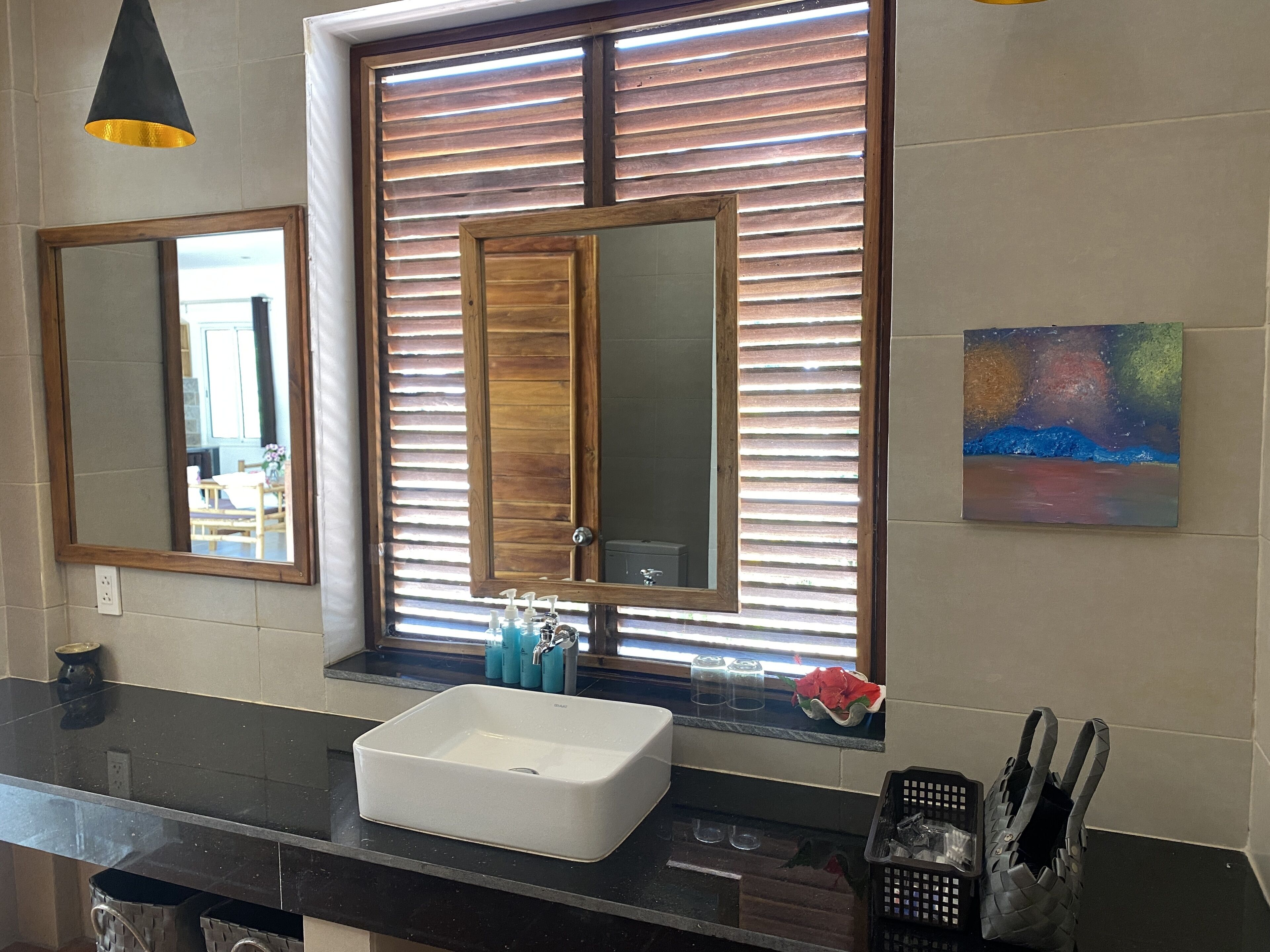 Une salle de bain avec un grand miroir, un lavabo blanc, des stores en bois et un tableau accroché au mur.