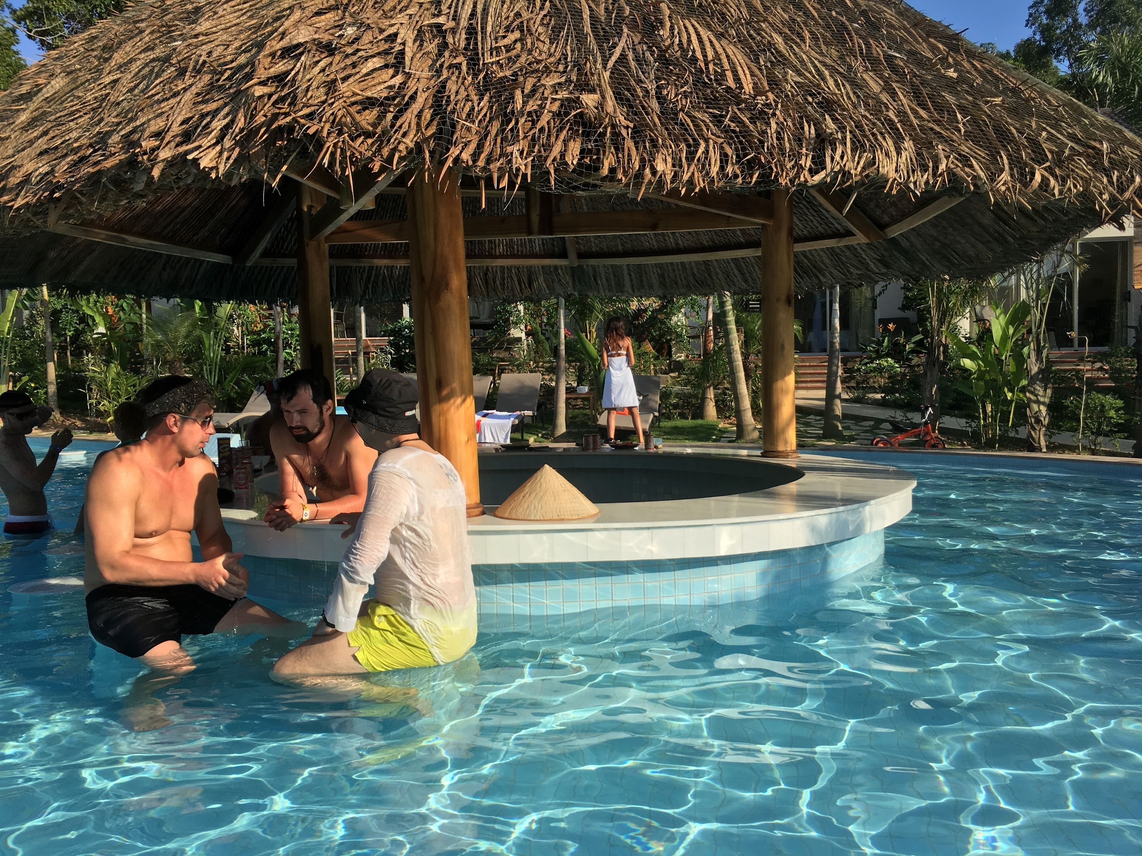 A pool area with a thatched roof structure, surrounded by palm trees and lounge chairs.