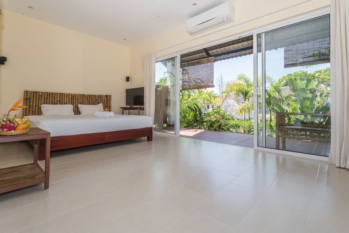 A spacious bedroom with a large bed, a bedside table with fruit, a TV, and a sliding glass door leading to a balcony with greenery.