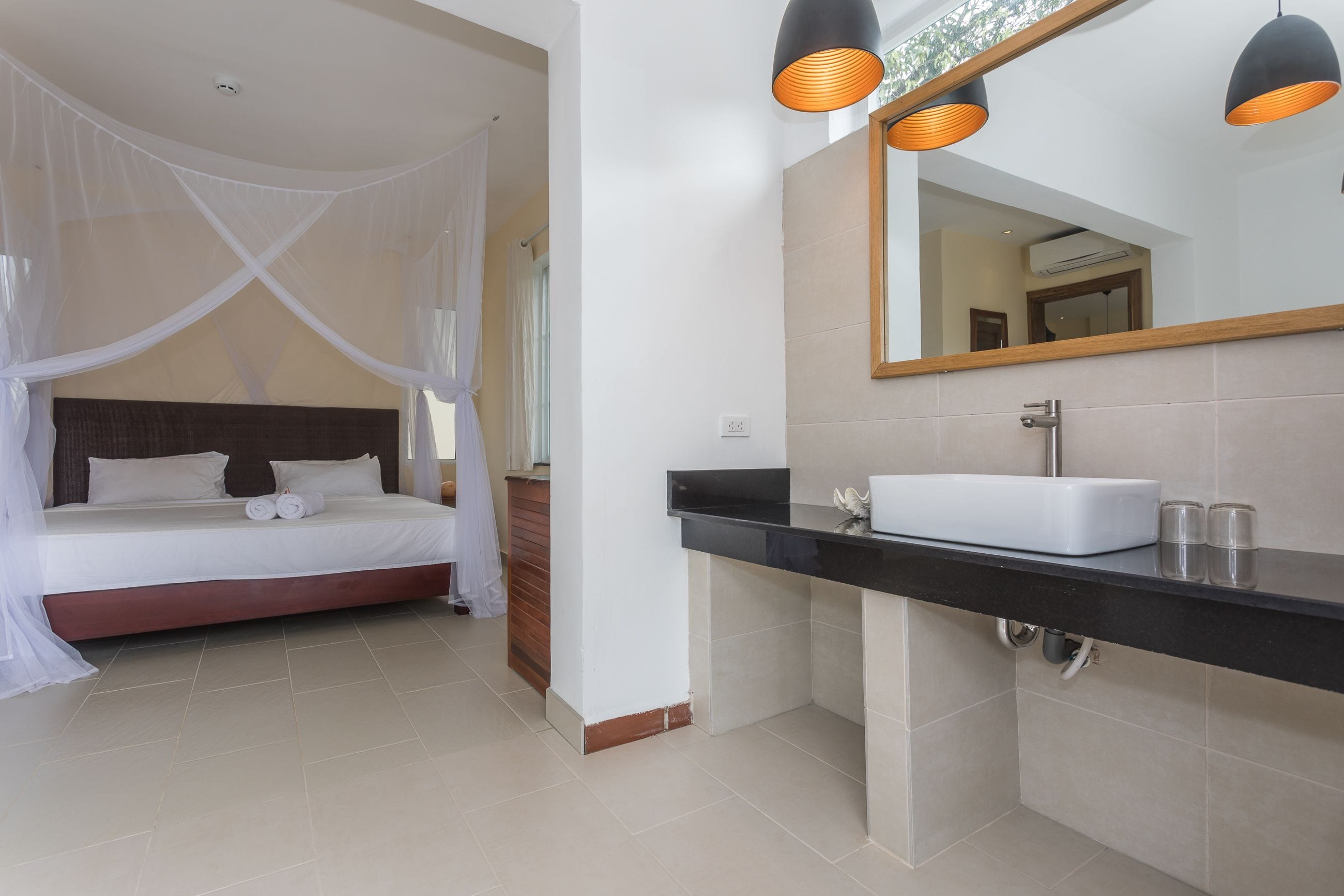 A bedroom with a canopy bed, a bathroom with a black countertop and a mirror, and a tiled floor.