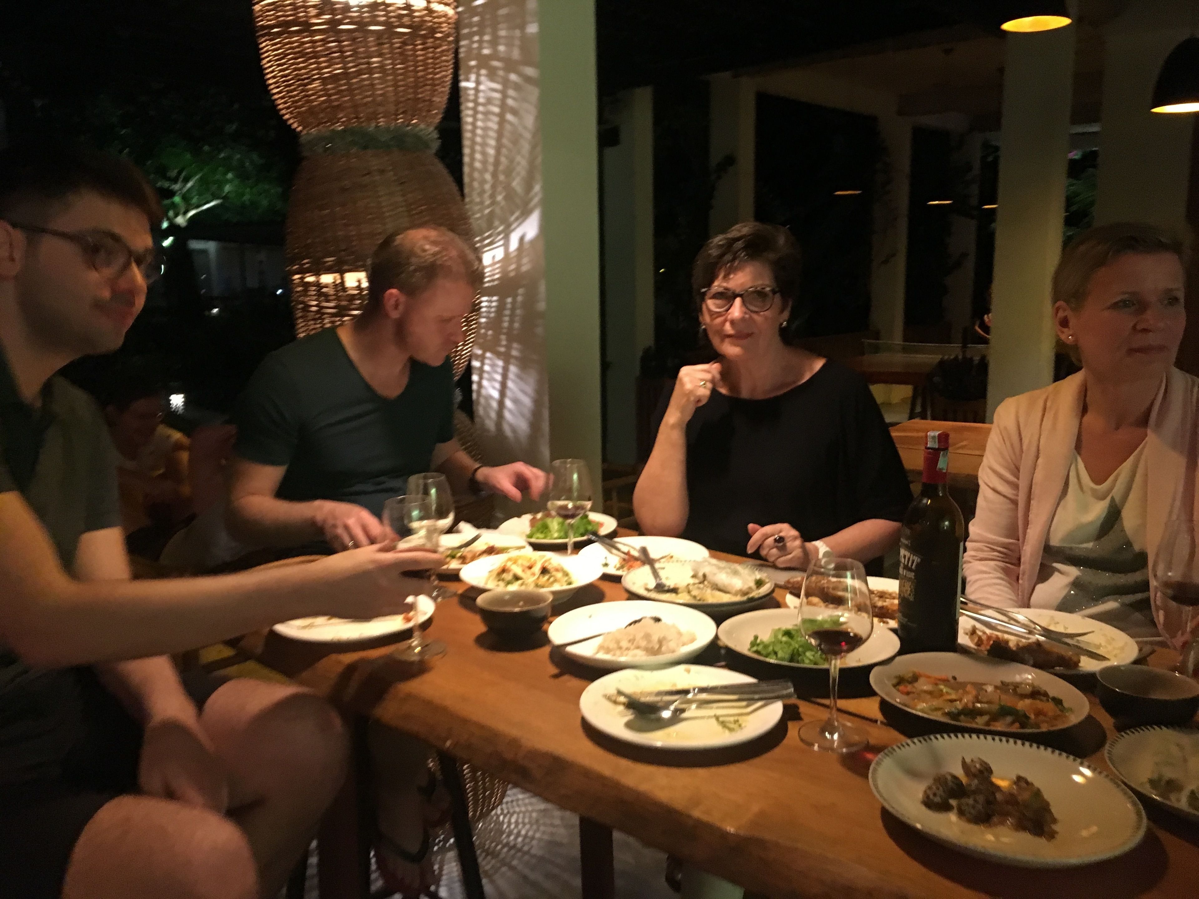 Un grupo de personas cenando en un restaurante con una mesa de madera, varios platos y copas de vino.