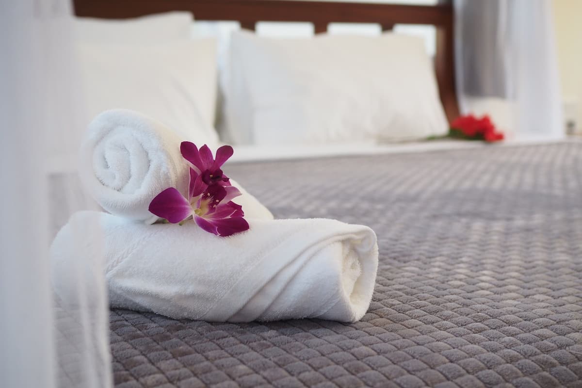 A neatly made bed with a rolled white towel and a purple flower on the bedspread.
