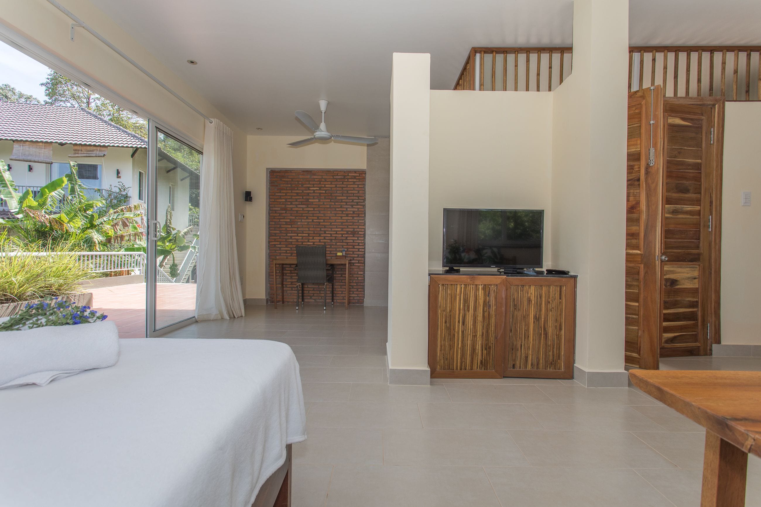 A hotel room with a bed, a TV, a wooden cabinet, a ceiling fan, and a balcony with plants.