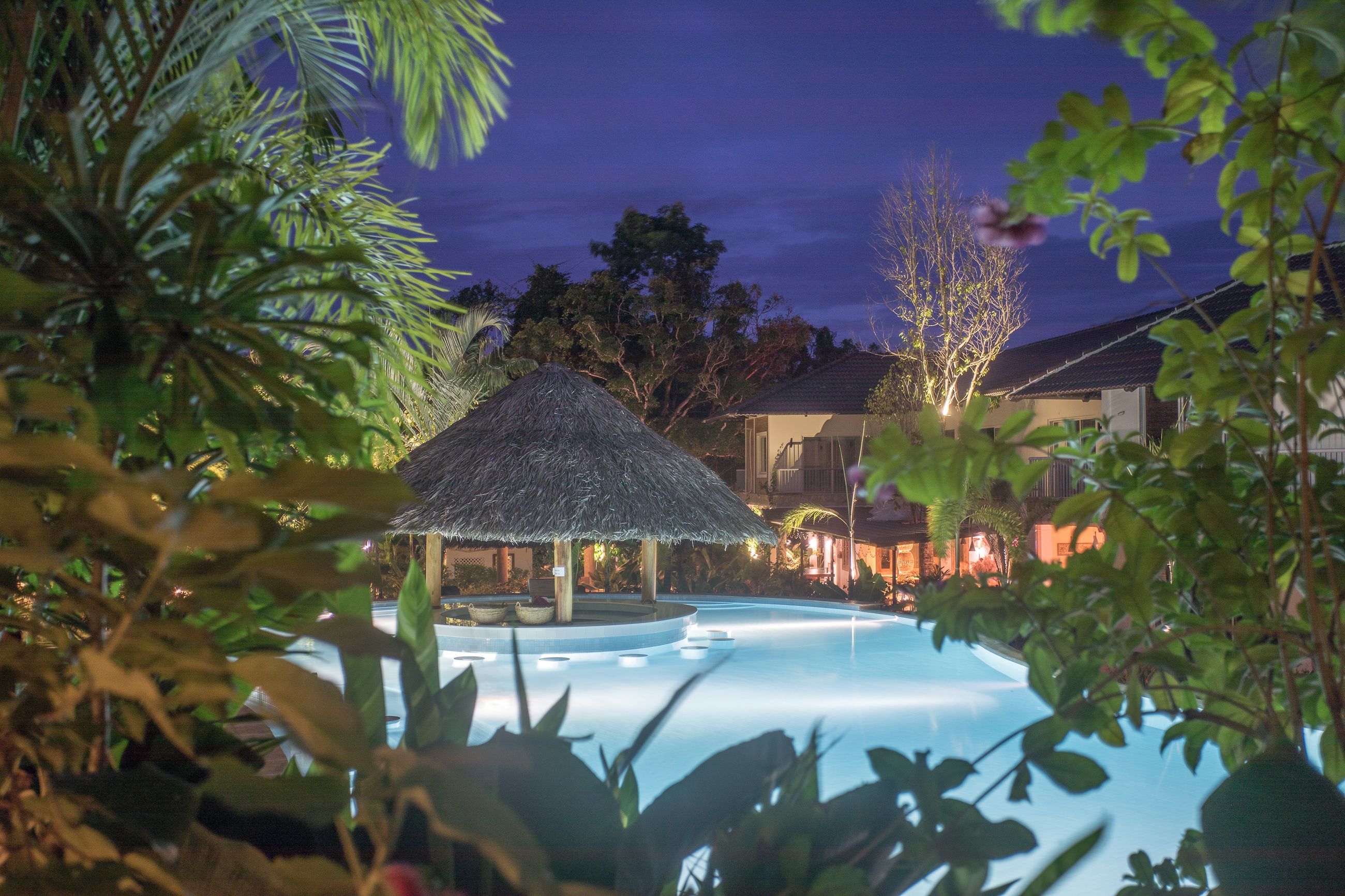 Un área de piscina con una estructura de techo de paja, rodeada de exuberante vegetación y un edificio con iluminación cálida.