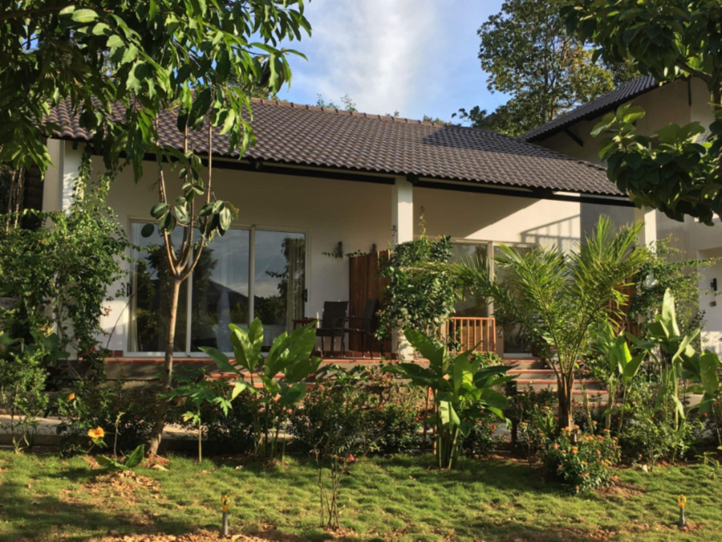 Una casa con techo de tejas, grandes ventanales y un jardín con diversas plantas y flores.