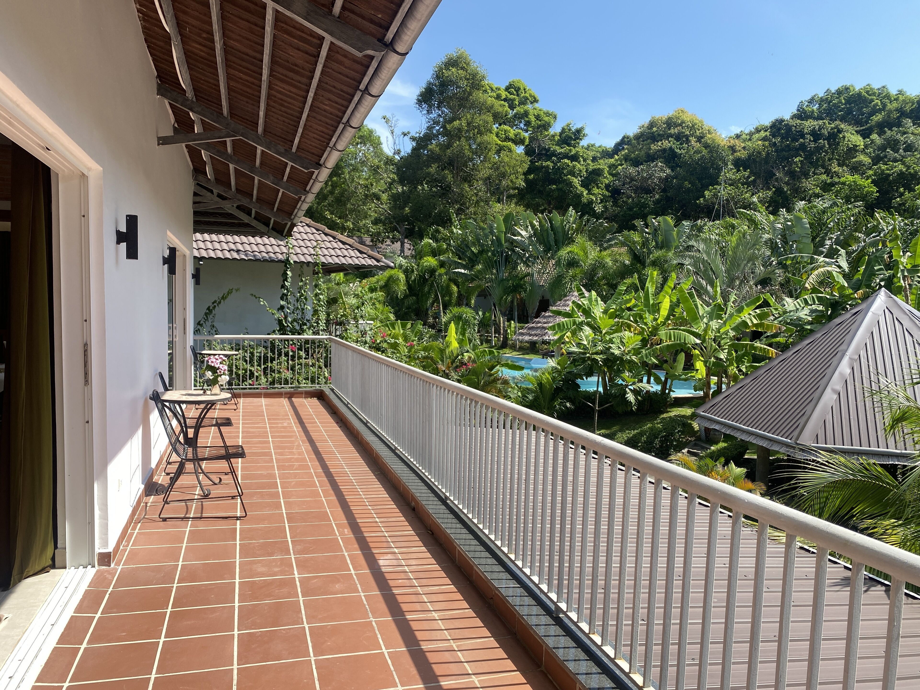 A balcony with a tiled floor, a metal railing, and a view of a lush garden and a pool.