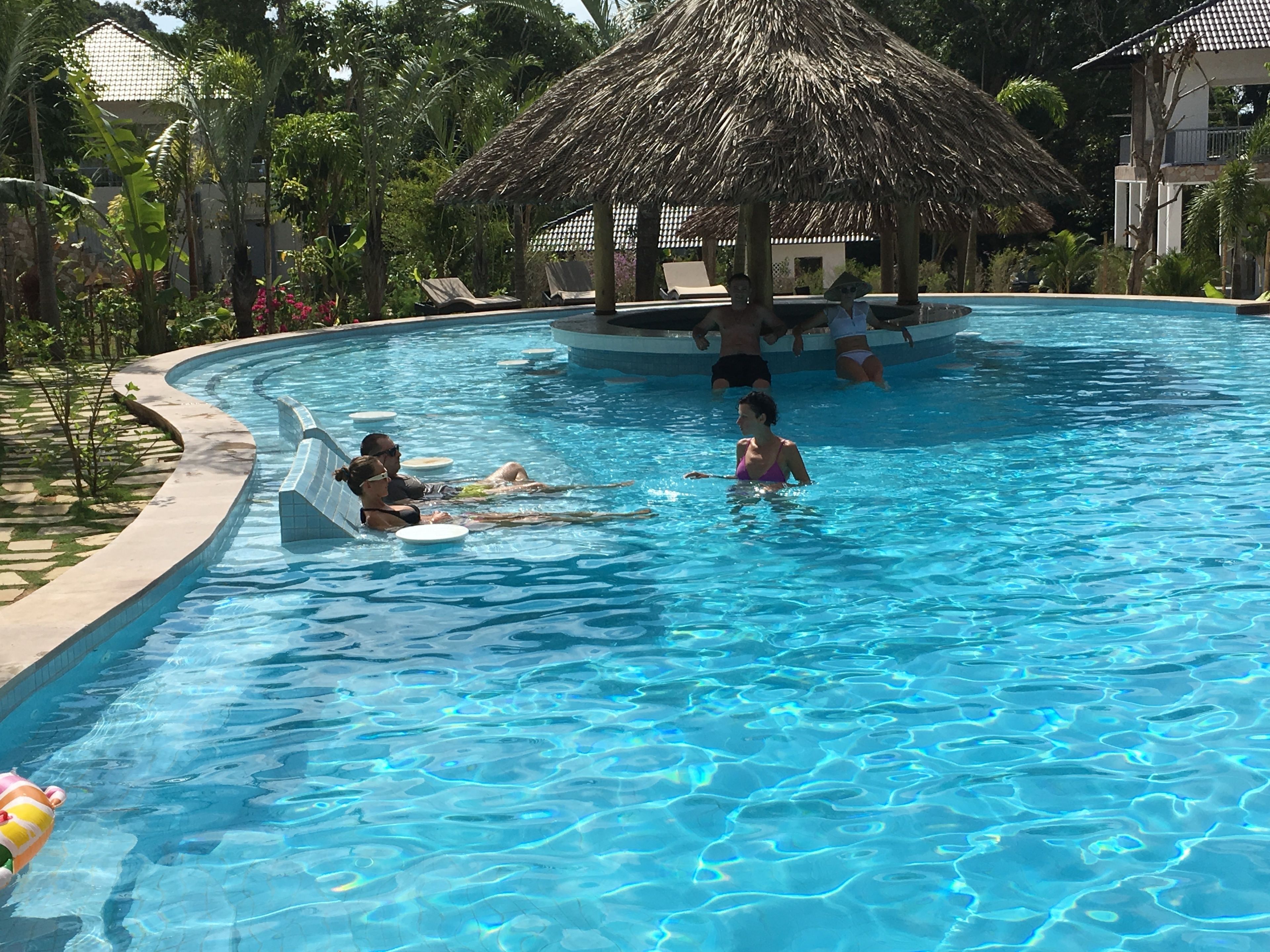 Una piscina con gente relajándose en sillones de descanso y flotadores, rodeada de plantas tropicales y una estructura con techo de paja.