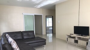 Un salon moderne avec un canapé d’angle, un téléviseur à écran plat posé sur un meuble en bois et une porte-fenêtre coulissante donnant sur une autre pièce.
