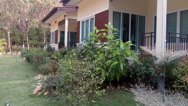 Une maison à deux étages dotée d’un balcon, entourée d’un jardin bien entretenu où poussent différentes espèces de plantes et où s’étend une pelouse.