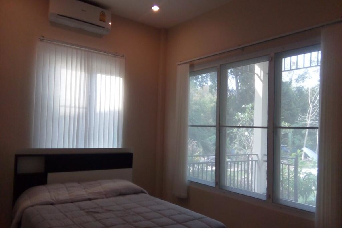 A bedroom with a bed, an air conditioner, a window with blinds, and a view of the outdoors.