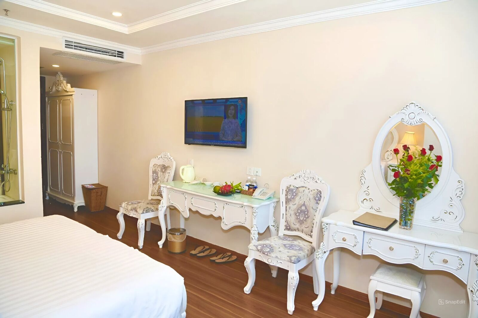 Habitación de hotel con cama, escritorio con teléfono, silla, tocador con espejo y un jarrón con flores.