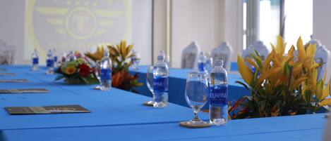 Un salón de conferencias con mantel azul, botellas de agua y una pantalla de proyector mostrando un logotipo.