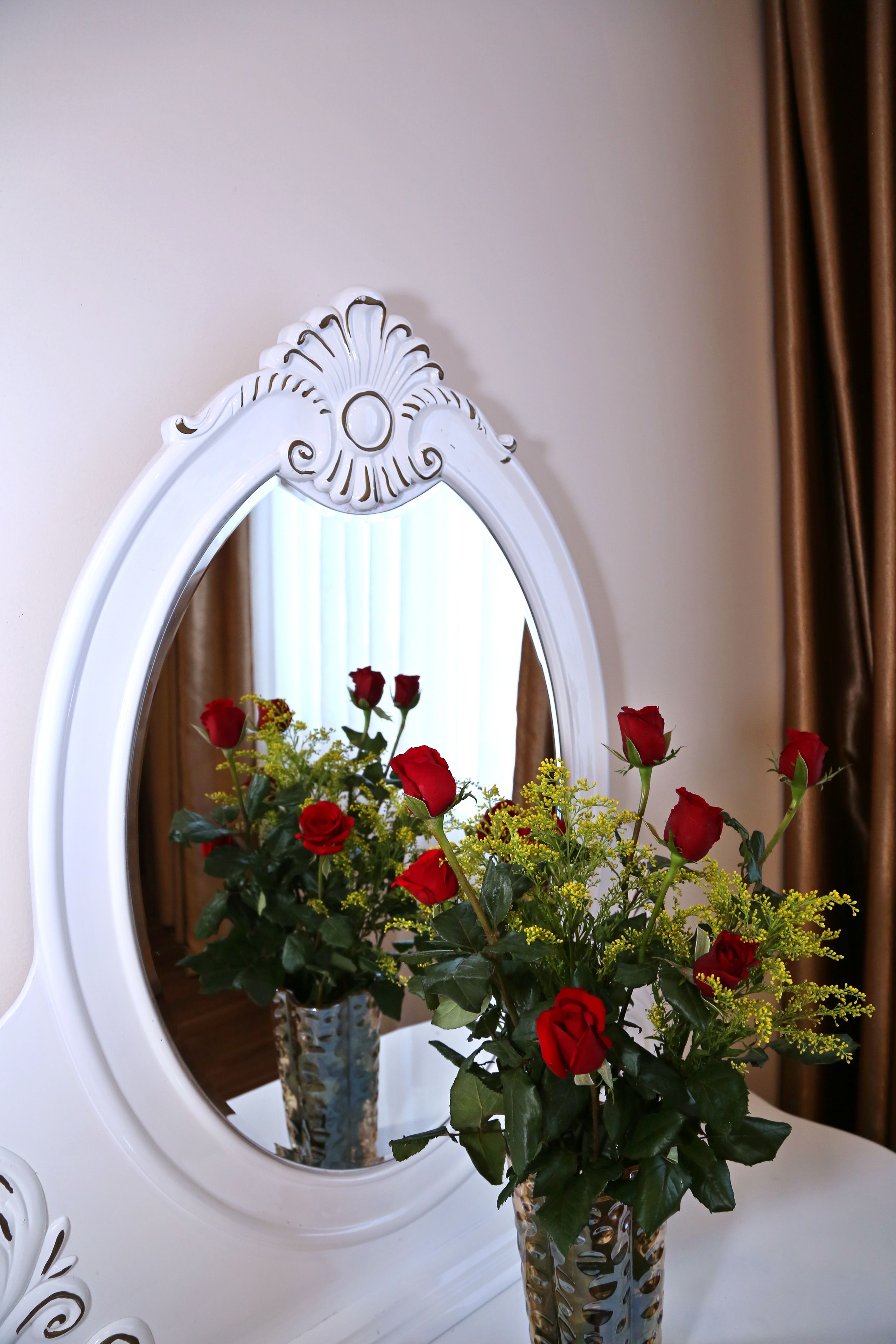 A mirror with an ornate frame reflecting a vase with red roses and greenery.