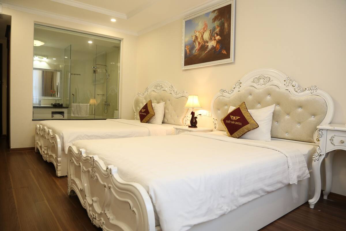 A hotel room with two large beds, a glass shower enclosure, a painting on the wall, and a nightstand with a lamp.