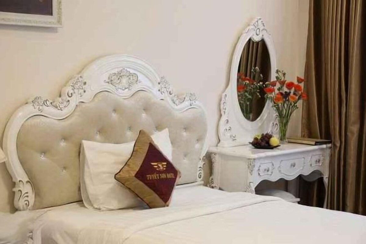 Una camera d'albergo con testiera capitonata, uno specchio, un comodino con un vaso di fiori e un cuscino decorativo.