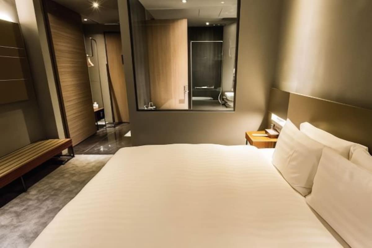 Una habitación de hotel moderna con una cama grande, un banco de madera, una mesita de noche y un espejo que refleja el baño.