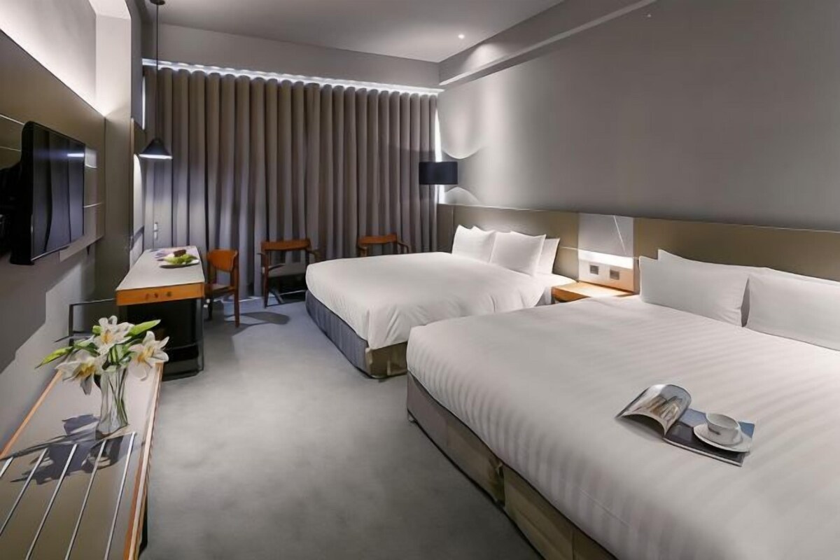 A hotel room with two beds, a desk with a chair, a TV, and a large window with curtains.