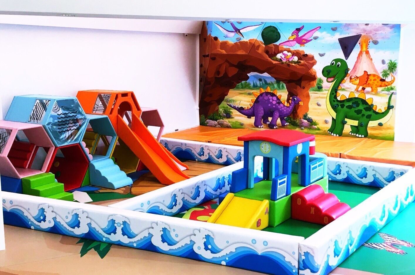 Zona de juegos infantiles con diversas estructuras coloridas, que incluyen toboganes y elementos para escalar, con un mural de fondo que muestra dinosaurios y un volcán.