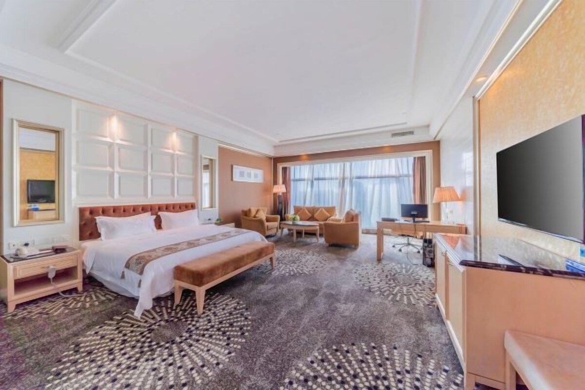A hotel room with a large bed, a desk, a chair, a television, and a large window.